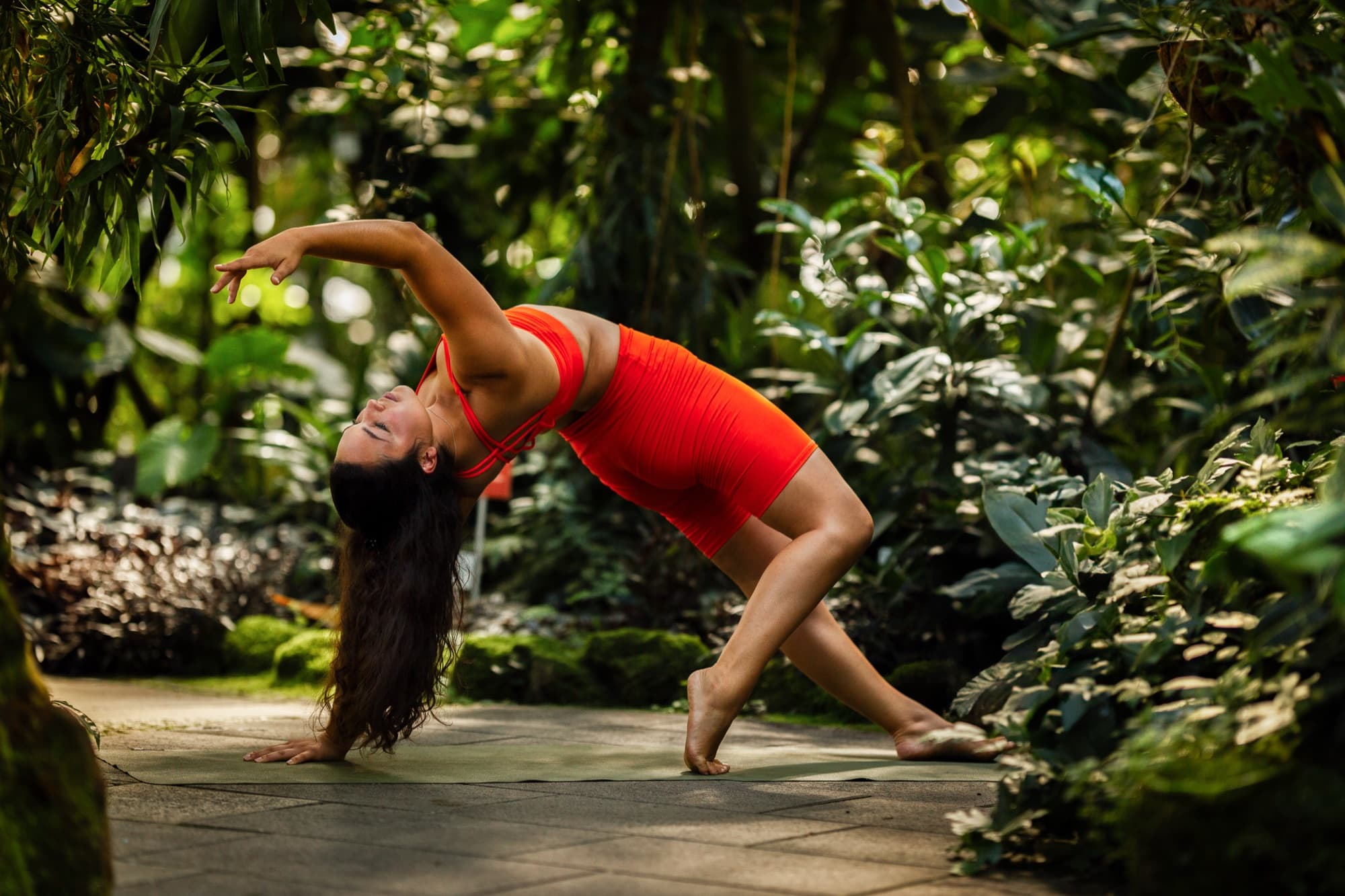 Sophie in einer dynamischen Yoga-Pose im Gewächshaus, orange Yoga-Outfit