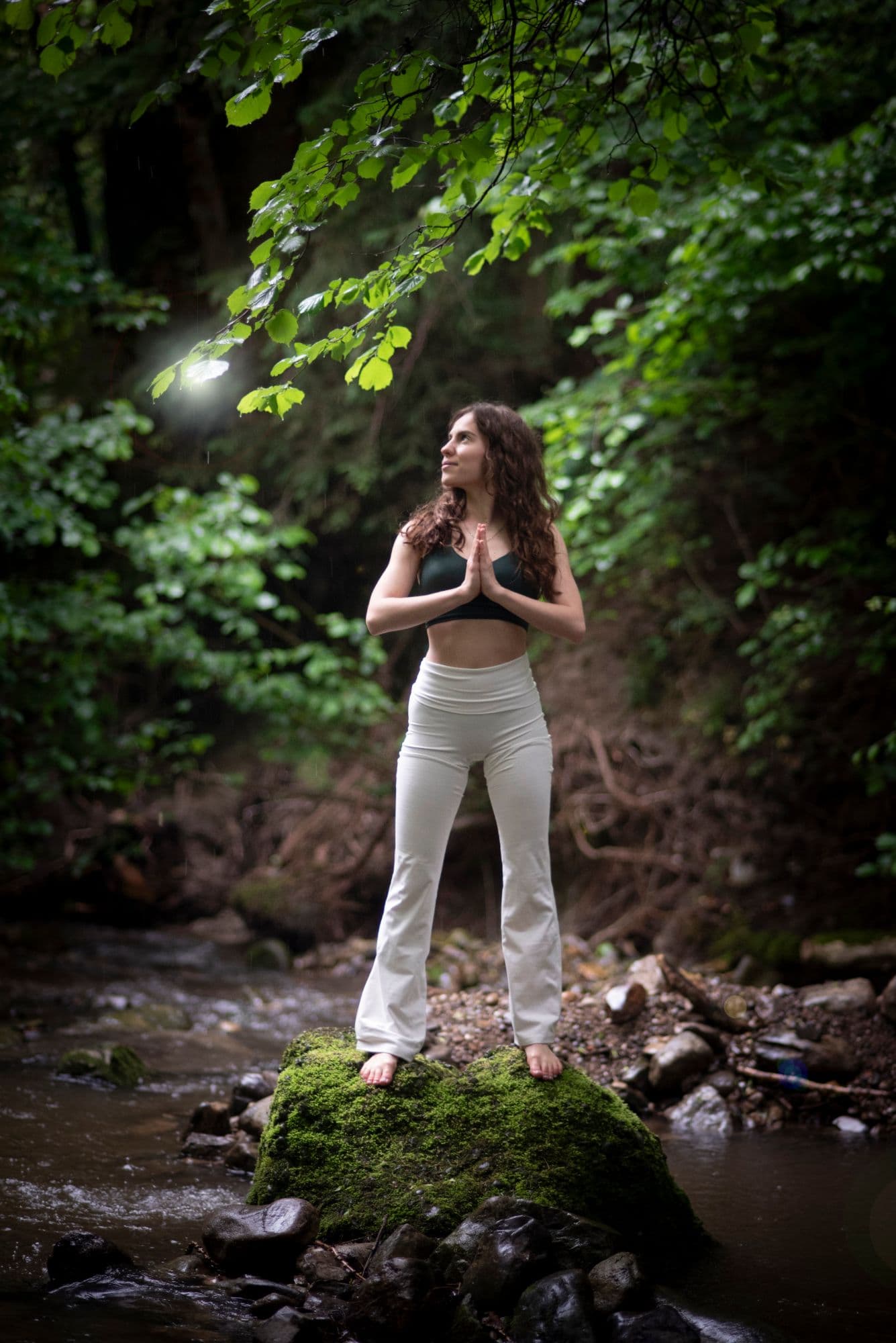 Yogalehrerin Julia Korel steht barfuß auf einem moosbewachsenen Stein an einem Waldbach, die Hände vor der Brust zusammengelegt